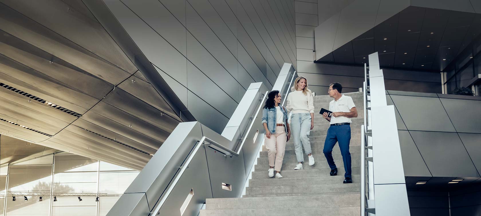 Two women and a BMW Welt employee walk down the stairs of BMW Welt during factory pickup