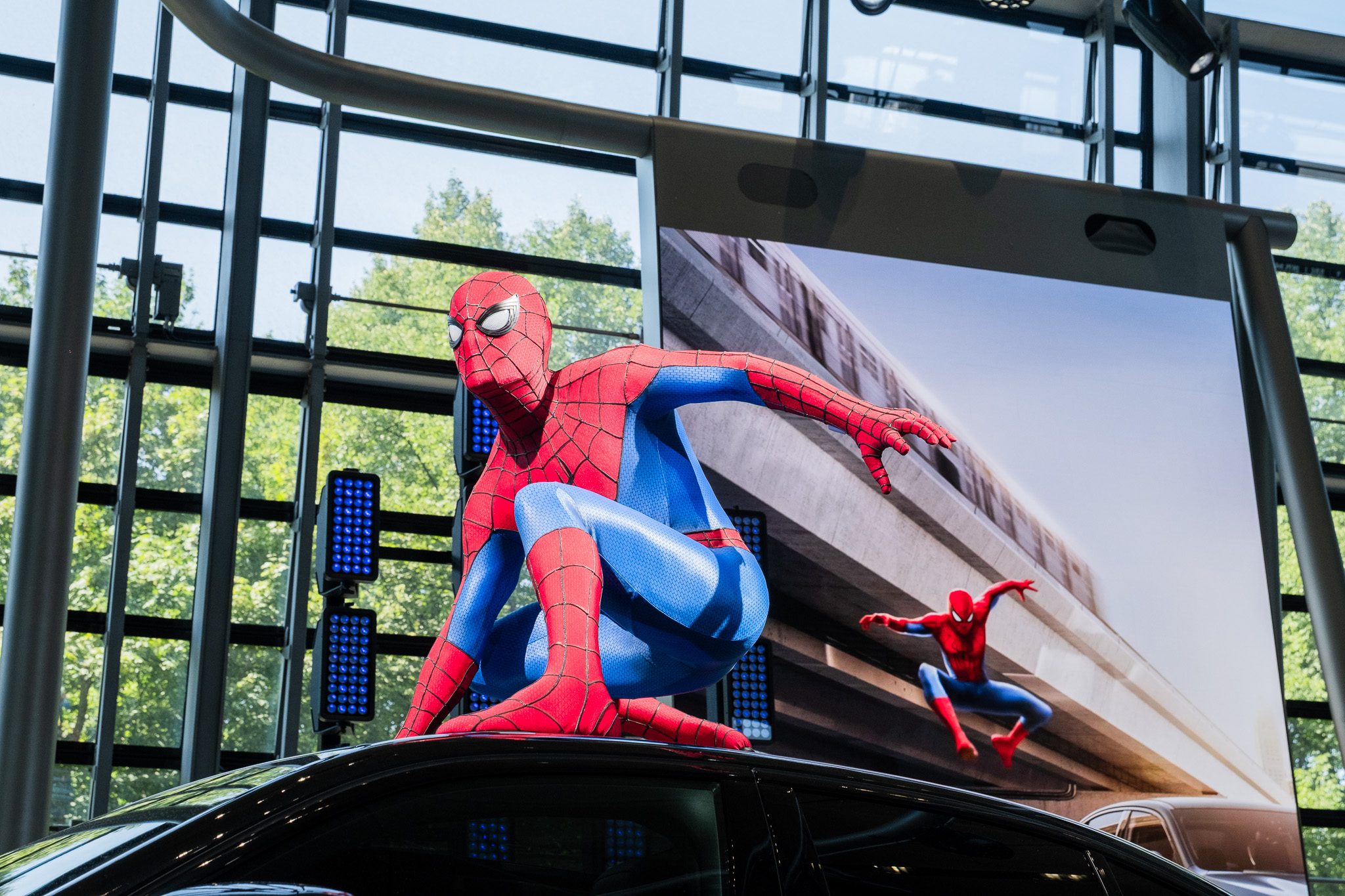 Black BMW 5 sedan displayed at BMW Welt with a Spider-Man figure on the vehicle roof in front of a large Spider-Man themed backdrop.
