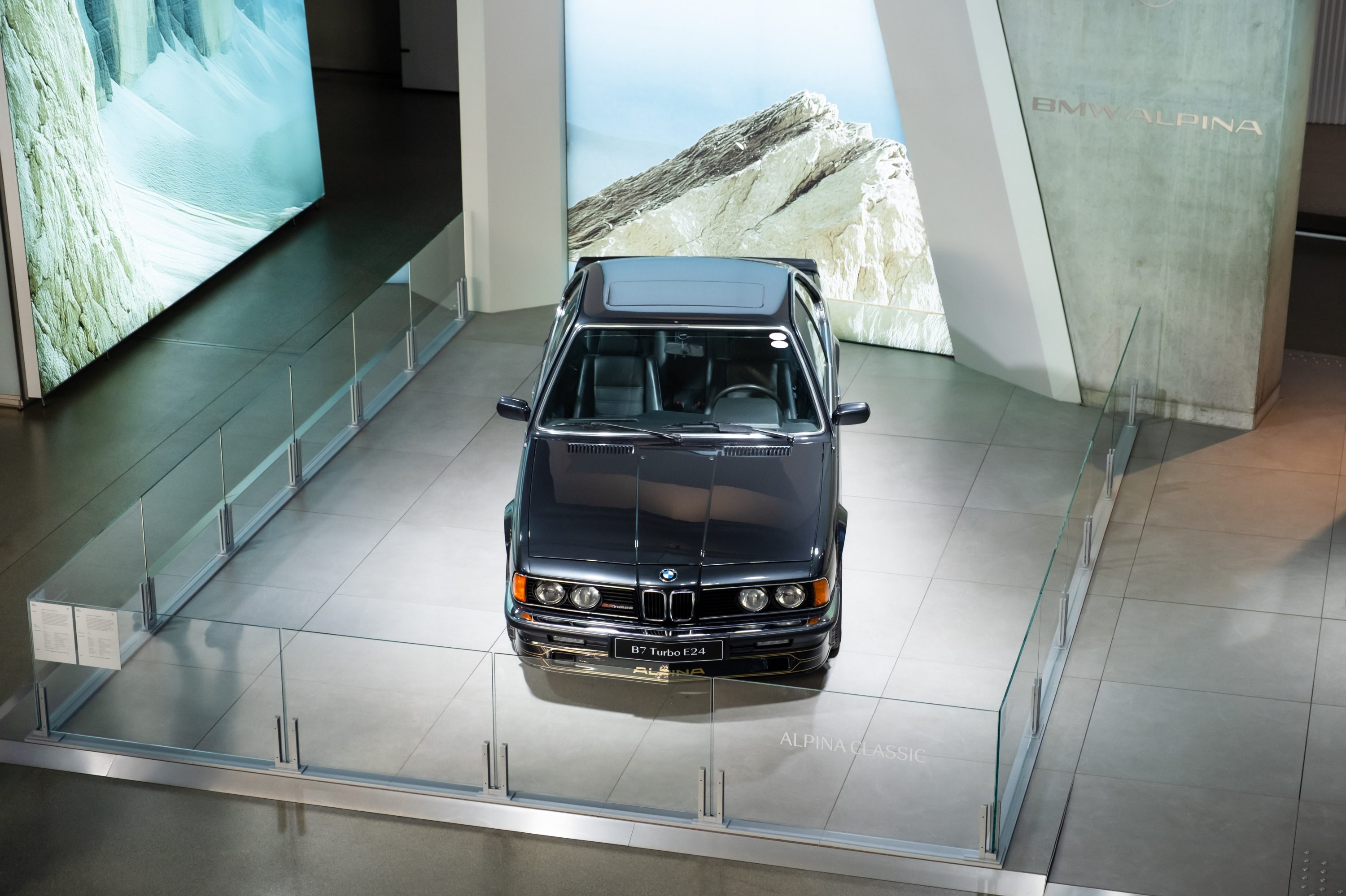 Alpina B7 Turbo (E24) displayed at BMW Welt on a glass-enclosed platform in a modern exhibition space with illuminated mountain landscape panels.